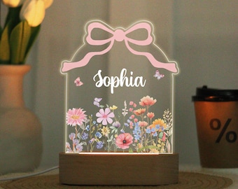Pode incluir: Luz noturna personalizada com o nome "Sophia" iluminado em branco. O painel de acrílico apresenta um design de laço rosa e um jardim floral colorido. A base é de madeira. Borboletas também estão incluídas no design.