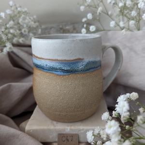 May include: A handcrafted ceramic mug with a distinctive design. The top section is white, flowing into a blue and brown band, and then a textured tan base. The mug has a curved handle and is set on a stone block.