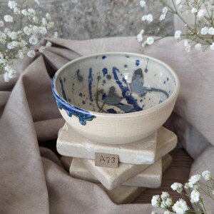 May include: A cream-coloured ceramic bowl with a blue-splattered interior design and a dark blue drip pattern on the rim. It rests on a stack of light-coloured square stone blocks. The tag on the blocks reads "A73".