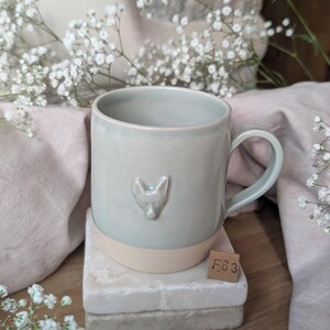 May include: A light sage green ceramic mug with a smooth glaze and a small, raised fox head relief on the front. The base of the mug is unglazed, showing a matte beige clay finish. A small square tag with the text F6 3 is placed near the base.