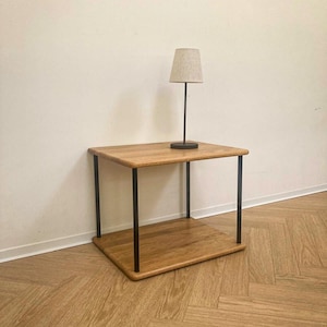 Può includere: Un tavolino a due livelli con due ripiani rettangolari in legno marrone chiaro sostenuti da quattro sottili gambe in metallo nero. Una piccola lampada da tavolo con una base sottile in metallo nero e un paralume in tessuto beige è appoggiata.