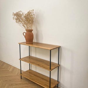 Op de afbeelding: Een rek met drie niveaus, voorzien van lichtbruine houten planken en een zwart metalen frame. Een terracotta-kleurige keramische kan met een boeket gedroogde beige bloemen staat op de bovenste plank van het rek tegen een witte muur.