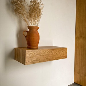 Peut inclure: Une étagère flottante rectangulaire en bois clair avec un motif de grain visible. Une cruche en céramique de couleur terre cuite est posée sur l'étagère, contenant un grand arrangement de tiges botaniques séchées brun clair.