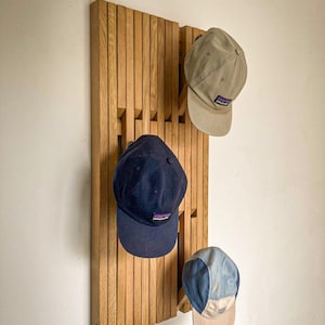 Könnte beinhalten: Ein vertikales Wandregal aus hellen Holzlatten mit drei hervorstehenden Haken. An den Haken hängen drei Baseballkappen: eine beige Kappe, eine dunkelmarineblaue Kappe und eine mehrfarbige blau-beige Kappe, die jeweils mit einem kleinen Logo versehen sind.