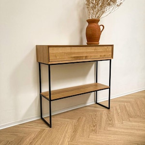 Peut inclure: Une console rectangulaire en bois dotée d'un tiroir unique et d'une étagère inférieure, soutenue par un cadre en métal noir. Un vase en céramique de couleur terre cuite contenant des fleurs séchées est posé sur le dessus de la console.