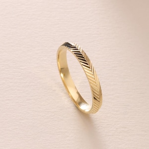 May include: A gold ring with a chevron pattern. The ring is set against a neutral background with a soft, warm light. The ring is a simple band with a textured surface.