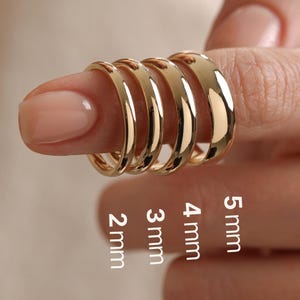 May include: Four polished gold-tone bands with smooth, rounded surfaces. The rings are arranged in a row, with text labels indicating widths of 2 mm, 3 mm, 4 mm, and 5 mm. Each ring features a simple, solid design with a reflective, metallic finish.