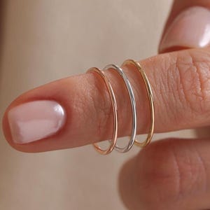 Könnte beinhalten: Drei dünne, glatte Metallringe, die zusammen an einem Finger getragen werden. Das Set besteht aus einem roségoldfarbenen Ring, einem silberfarbenen Ring und einem gelbgoldfarbenen Ring. Jeder Ring hat ein schlichtes, rundes Design mit polierter Oberfläche.