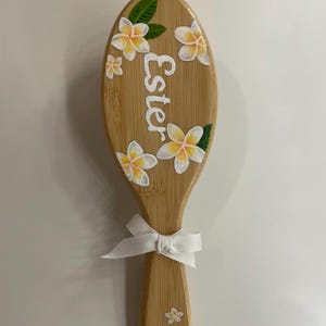 May include: A wooden hairbrush with the name "Ester" in white script. The brush is decorated with white and yellow flowers and green leaves. A white ribbon is tied at the base of the handle.