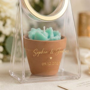 May include: A terracotta flower pot candle with a light blue succulent-shaped wax top, encased in a clear triangular frame with a gold rim. The pot features gold text reading Sophia & James, a small heart icon, and the date 09.12.2026 printed on it.