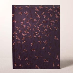 May include: A hardcover journal with a dark purple cover adorned with a pattern of flying birds in shades of purple and gold. The birds are depicted in various positions, creating a sense of movement and flight. The journal is rectangular and stands upright.