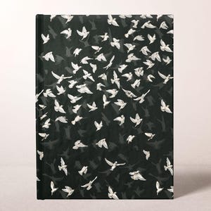 May include: A hardcover journal with a black cover featuring a pattern of white birds in flight. The birds are depicted in various sizes and positions, creating a dynamic and artistic design. The journal has a clean, minimalist aesthetic.