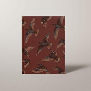 May include: A hardcover journal with a burgundy cover featuring a repeating pattern of birds in flight. The birds are depicted in shades of brown and tan, with outstretched wings against the solid background. The journal is rectangular.