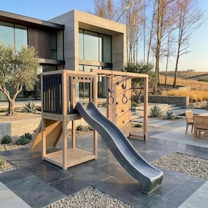 May include: A wooden outdoor playset with a gray slide, climbing wall, and monkey bars. The playset is set on a stone patio with a modern house in the background. The playset is surrounded by landscaping.