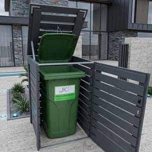 Op de afbeelding: Een donkergrijze buitenafvalbak met een groene afvalbak erin. De deksel van de bak staat open. De behuizing heeft een lamellenontwerp en een scharnierende deur. De achtergrond toont een modern huis met steen en glas.