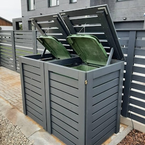 Puede incluir: Contenedor de basura exterior de dos compartimentos con diseño de listones gris oscuro. Las tapas están abiertas, revelando cubos interiores verdes. El contenedor está sobre una superficie pavimentada.