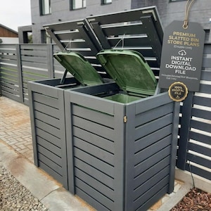 Op de afbeelding: Een dubbele kliko-ombouw van donkergrijs hout met twee open deksels die groene kunststof afvalcontainers onthullen. Een hangend label toont de tekst: PREMIUM SLATTED BIN STORE PLANS (Double Unit) INSTANT DIGITAL DOWNLOAD FILE.