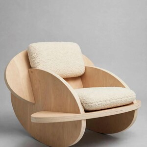 Può includere: Una poltrona realizzata in compensato di colore chiaro con un design a telaio circolare curvo. La seduta e lo schienale presentano spessi cuscini color bianco sporco con una texture effetto lana che contrasta con la struttura in legno.