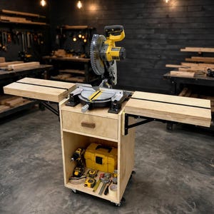 May include: A yellow and black miter saw mounted on a light-colored plywood mobile workstation. The unit features a central drawer, two fold-out side extensions, and an open lower shelf containing a drill, a yellow tool case, and various hand tools.