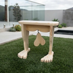 Könnte beinhalten: Ein Hocker aus hellem Holz mit einer flachen, rechteckigen Sitzfläche, die von zwei Beinen in Form menschlicher Unterschenkel getragen wird, komplett mit geschnitzten Füßen an der Basis und einem mittig hängenden herzförmigen Holzelement.