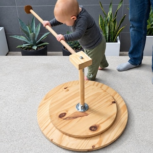 Könnte beinhalten: Ein hölzernes Balancierspielzeug bestehend aus zwei übereinanderliegenden, kreisförmigen Plattformen aus hellem Holz, die durch einen zentralen vertikalen Stab und eine horizontale Griffstange verbunden sind, gefertigt aus natürlichem Kiefernholz.