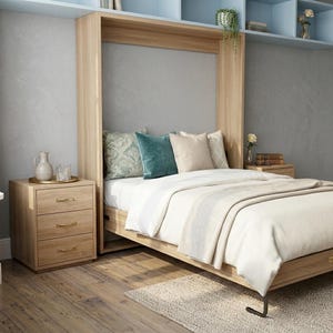 May include: A light wood Murphy bed frame with an integrated tall storage unit, featuring a white mattress, a textured white blanket, and four decorative pillows in shades of teal, beige, and patterned gray, next to a matching three-drawer nightstand.