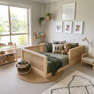 Op de afbeelding: Een lichthouten dagbed met rieten panelen, opgemaakt met olijfgroen beddengoed, een crèmekleurig kussen met drie boomafbeeldingen en een bruine teddybeer. De kamer bevat een houten boekenplank, een gevlochten mand, hangplanten en ingelijste kunst.