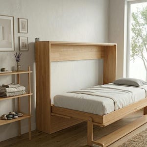 May include: A wooden wall bed in a bedroom. The bed is lowered, with a light-coloured mattress and pillow. A wooden shelving unit and framed artwork are visible on the left. The bed is made of light-coloured wood.