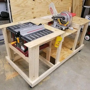 May include: A wooden workbench on casters featuring a built-in table saw and a miter saw. The table saw has a black and red base with a clear guard. The miter saw is silver and red with a yellow blade. A yellow cup is attached to the frame.