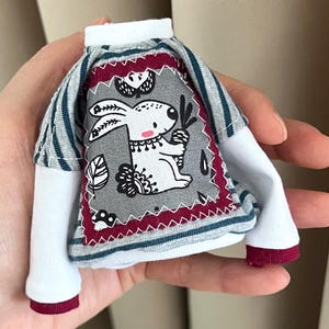 Könnte beinhalten: Ein kleines Puppenhemd mit weißem Körper und gestreiften Ärmeln. Die Vorderseite hat einen grauen Aufnäher mit einer weißen Kaninchenillustration, umrahmt von einem bordeauxroten Rand. Die Ärmel und Manschetten haben bordeauxrote Akzente.