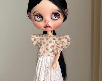 May include: A Blythe doll with black hair and large, expressive eyes. The doll wears a beige top with a pink floral pattern and a white eyelet skirt. Black shoes complete the outfit. The doll is held by a hand.