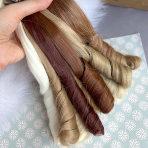 May include: A hand holds a variety of hair extensions in shades of blonde, brown, and auburn. The extensions are bundled and curled, showcasing different textures and colors. The background includes a white fluffy surface and a patterned paper.