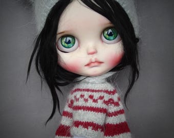 May include: A doll with black hair, large green eyes, and rosy cheeks, wearing a light gray knit hat with cat ears, a light gray and red striped long-sleeve sweater, and a white skirt with a subtle eyelet pattern on a plain gray background.