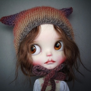 Könnte beinhalten: Eine Puppe mit großen, ausdrucksstarken braunen Augen und rosigen Wangen. Die Puppe trägt eine gestrickte Mütze in Braun-, Lila- und Orangetönen mit ohrenartigen Details. Ein passender Schal ist um den Hals gewickelt.