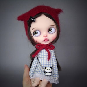 Può includere: Una bambola con capelli castano scuro indossa un berretto di lana rosso con orecchie da gatto e un vestito a maniche lunghe a quadri blu e bianchi con orlo in pizzo bianco. La bambola ha grandi occhi e un ciondolo a forma di panda sul petto.