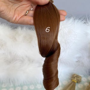 May include: A sample of brown hair extensions, shade number 6, held in a hand. The hair is twisted to show its texture and color. Other hair samples are visible in the background.