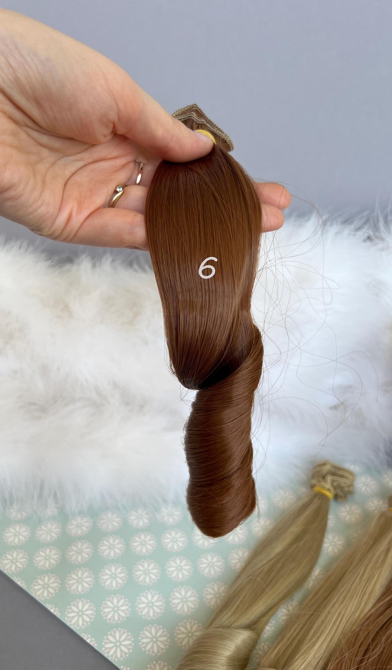 May include: A sample of brown hair extensions, shade number 6, held in a hand. The hair is twisted to show its texture and color. Other hair samples are visible in the background.
