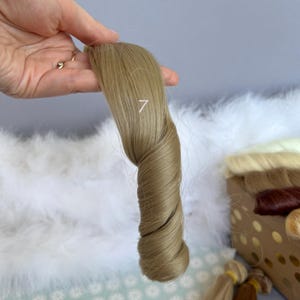 May include: A sample of light brown hair extensions, twisted to show the texture and color. The number "7" is printed on the hair. Other hair samples are visible in the background.