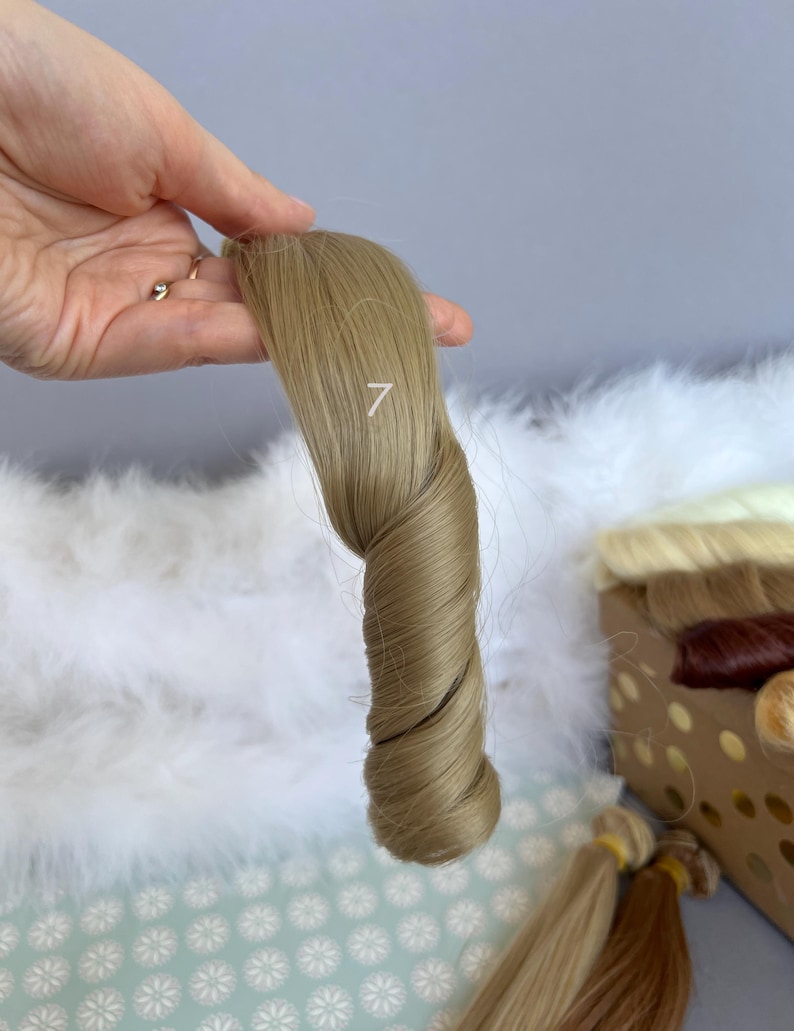 May include: A sample of light brown hair extensions, twisted to show the texture and color. The number "7" is printed on the hair. Other hair samples are visible in the background.