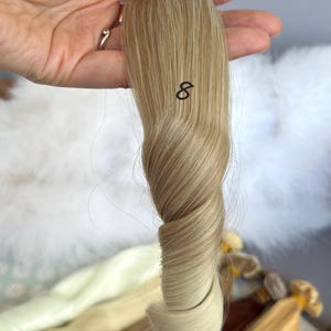 May include: A sample of blonde hair extensions, held in a hand, with the number 8 printed on the hair. The hair is a light blonde color with darker roots. Other hair samples are visible in the background.