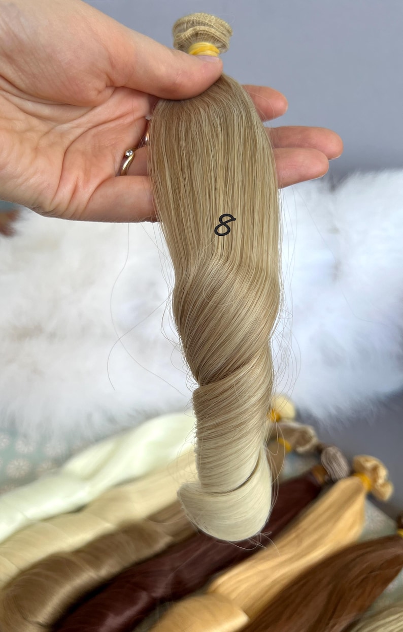 May include: A sample of blonde hair extensions, held in a hand, with the number 8 printed on the hair. The hair is a light blonde color with darker roots. Other hair samples are visible in the background.