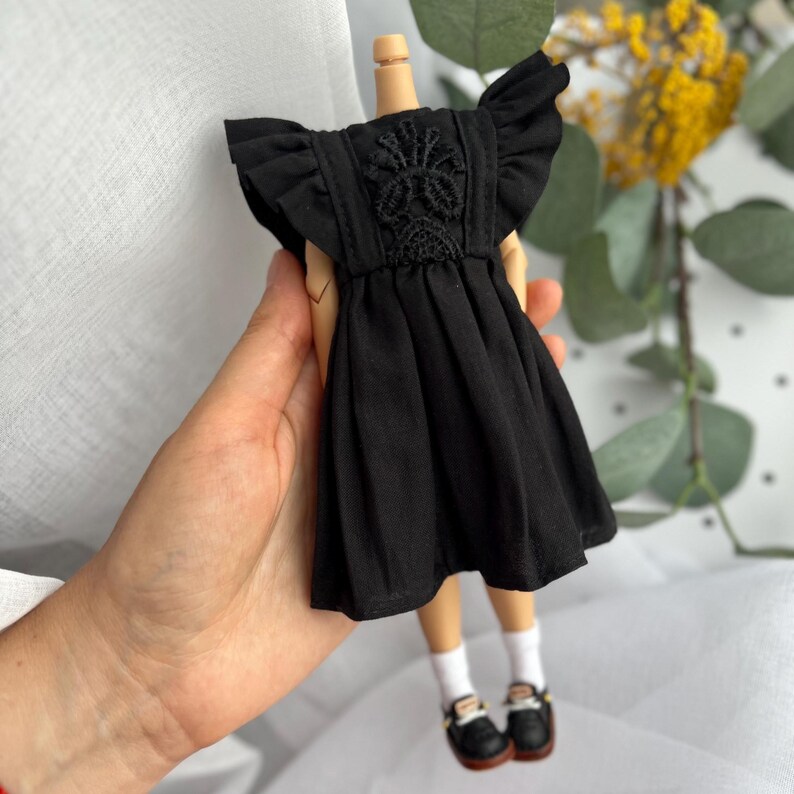 May include: A black doll dress featuring ruffled sleeves and a central panel with intricate black lace embroidery. The garment has a gathered skirt and is displayed on a doll with white socks and black shoes, showcasing detailed craftsmanship.