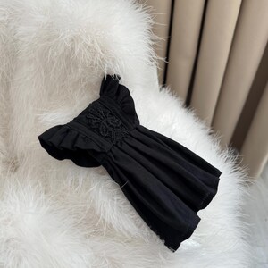 May include: A black top featuring short ruffled sleeves and a central panel of intricate floral lace embroidery. The garment has a gathered, pleated bodice that creates a flared silhouette, with a round neckline and a solid black fabric construction.