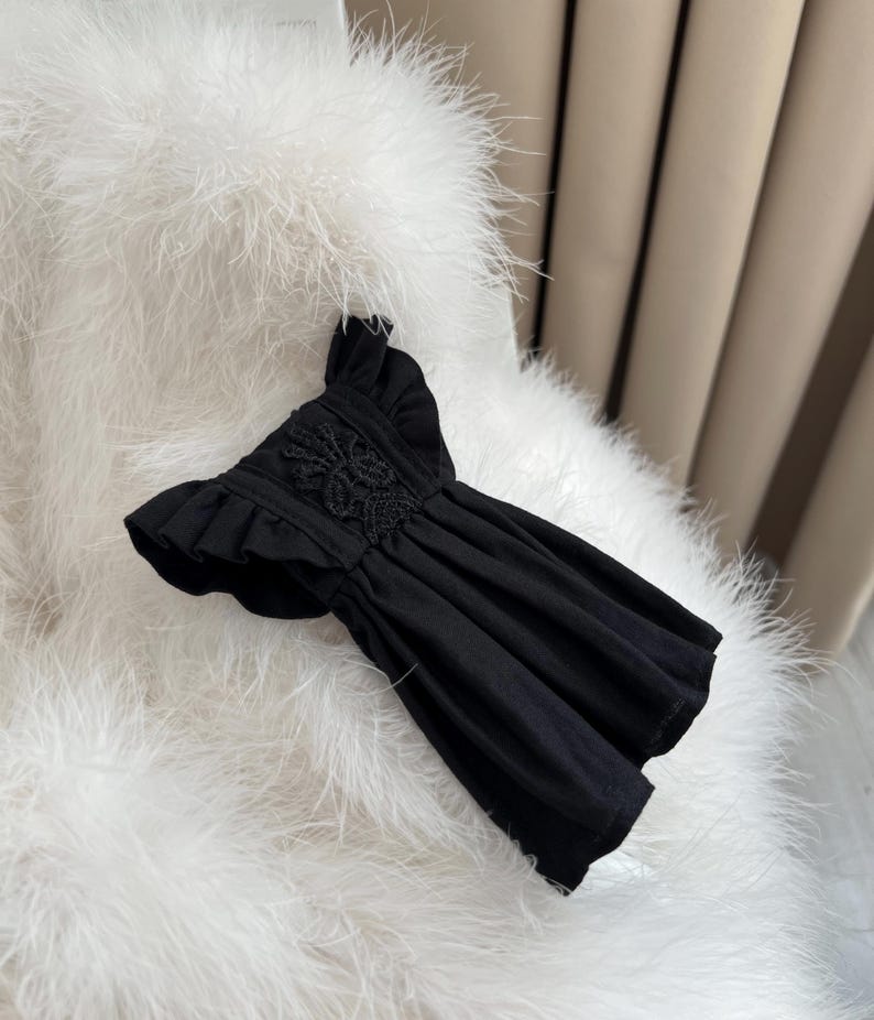 May include: A black top featuring short ruffled sleeves and a central panel of intricate floral lace embroidery. The garment has a gathered, pleated bodice that creates a flared silhouette, with a round neckline and a solid black fabric construction.