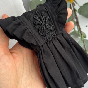 May include: A small black dress featuring ruffled sleeves and a central panel with intricate black floral embroidery. The garment has a pleated skirt section and is constructed from a solid black fabric with a consistent matte texture throughout.