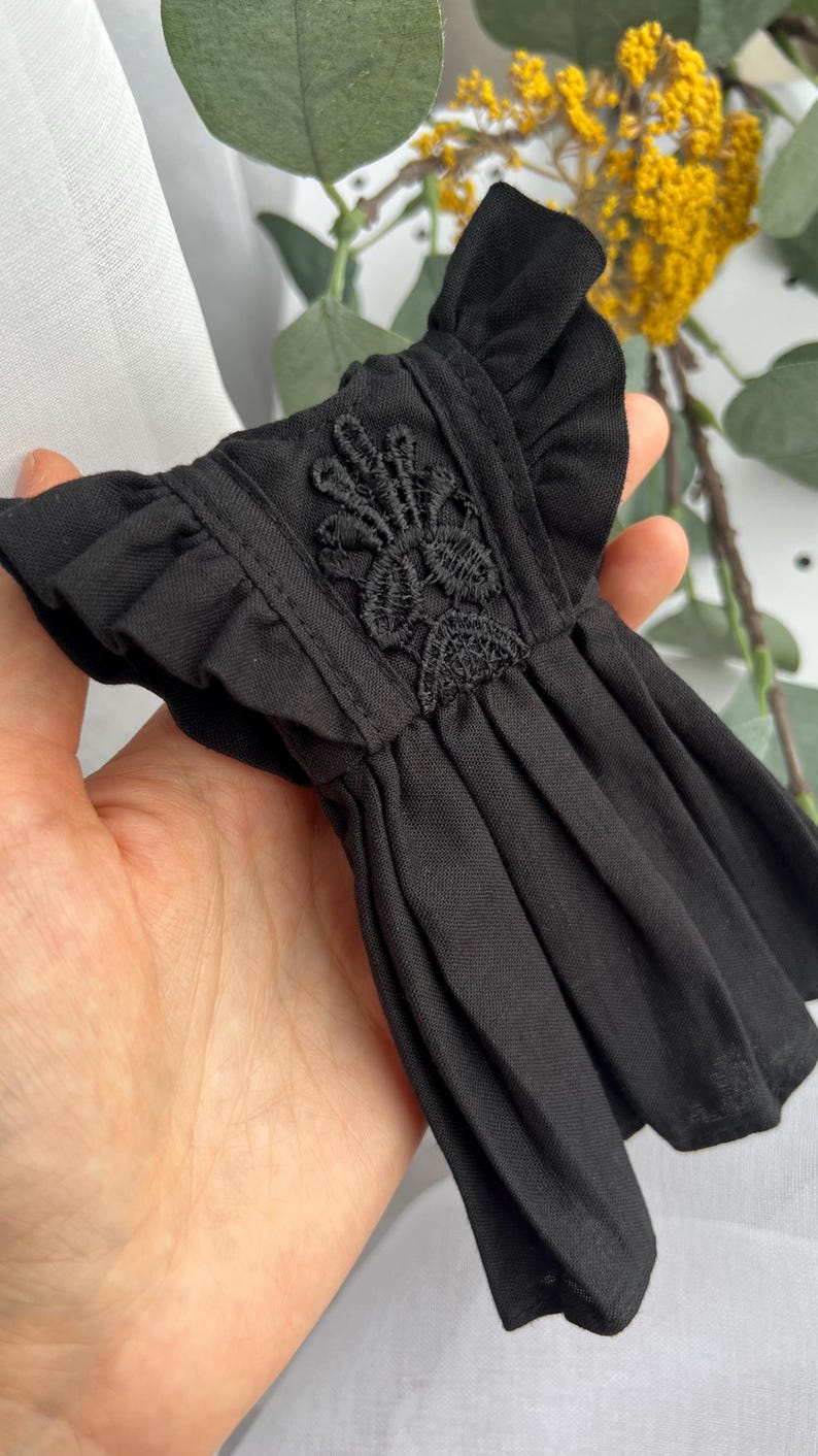 May include: A small black dress featuring ruffled sleeves and a central panel with intricate black floral embroidery. The garment has a pleated skirt section and is constructed from a solid black fabric with a consistent matte texture throughout.