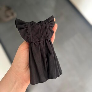 May include: A small black dress for a doll, featuring short ruffled sleeves, a gathered skirt, and vertical stitched detailing on the bodice. The garment is made of a solid black fabric and is designed with a simple, functional construction.