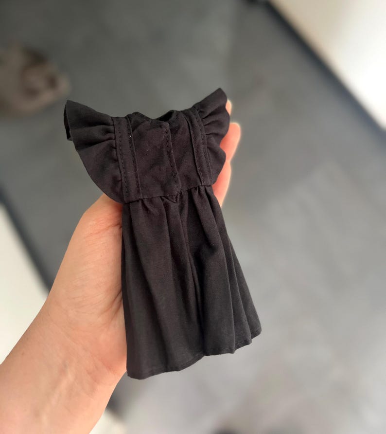 May include: A small black dress for a doll, featuring short ruffled sleeves, a gathered skirt, and vertical stitched detailing on the bodice. The garment is made of a solid black fabric and is designed with a simple, functional construction.