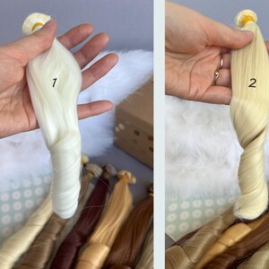 May include: Two bundles of doll hair in shades of white and blonde are held up for display. The hair is styled in a twisted, wavy pattern. Other bundles of hair in various shades of brown and blonde are visible in the background.