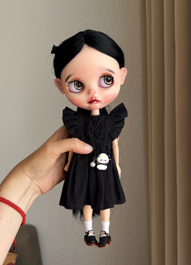 May include: A doll with pale skin, large brown eyes, and black hair styled in a centre part. The doll wears a black dress with ruffled sleeves, a small white panda-shaped crossbody bag with a pearl charm, white socks, and black shoes.